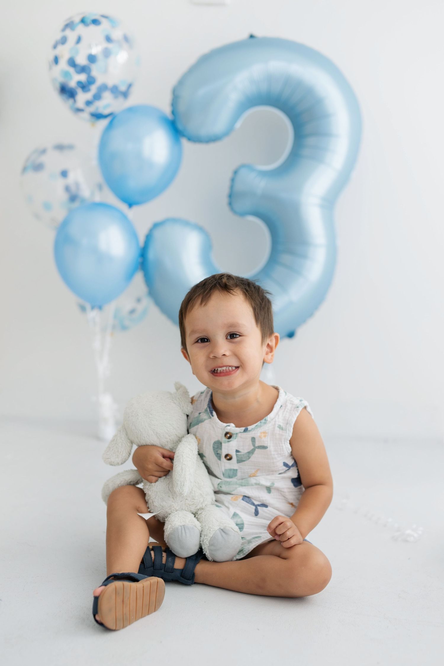 Smiling toddler boy sitting on white floor holding stuffed animal with blue number 3 birthday balloons behind him.
