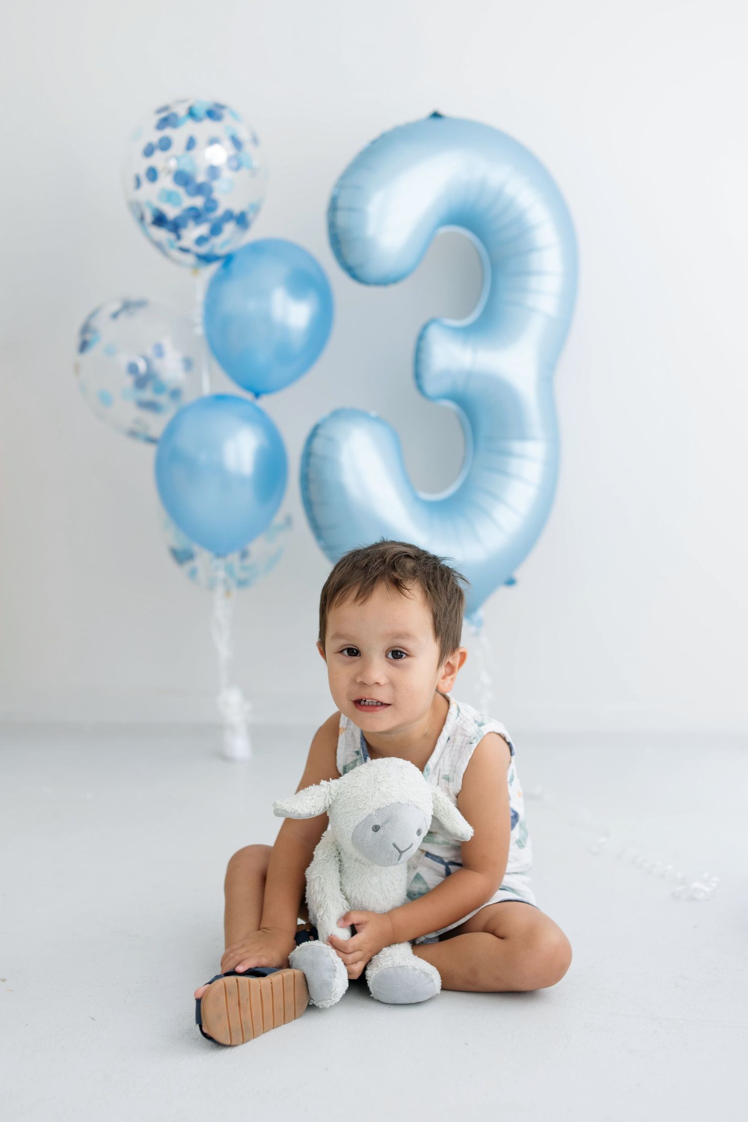 A toddler boy sits on a white floor holding a stuffed lamb toy, surrounded by blue balloons and a number 3 balloon.