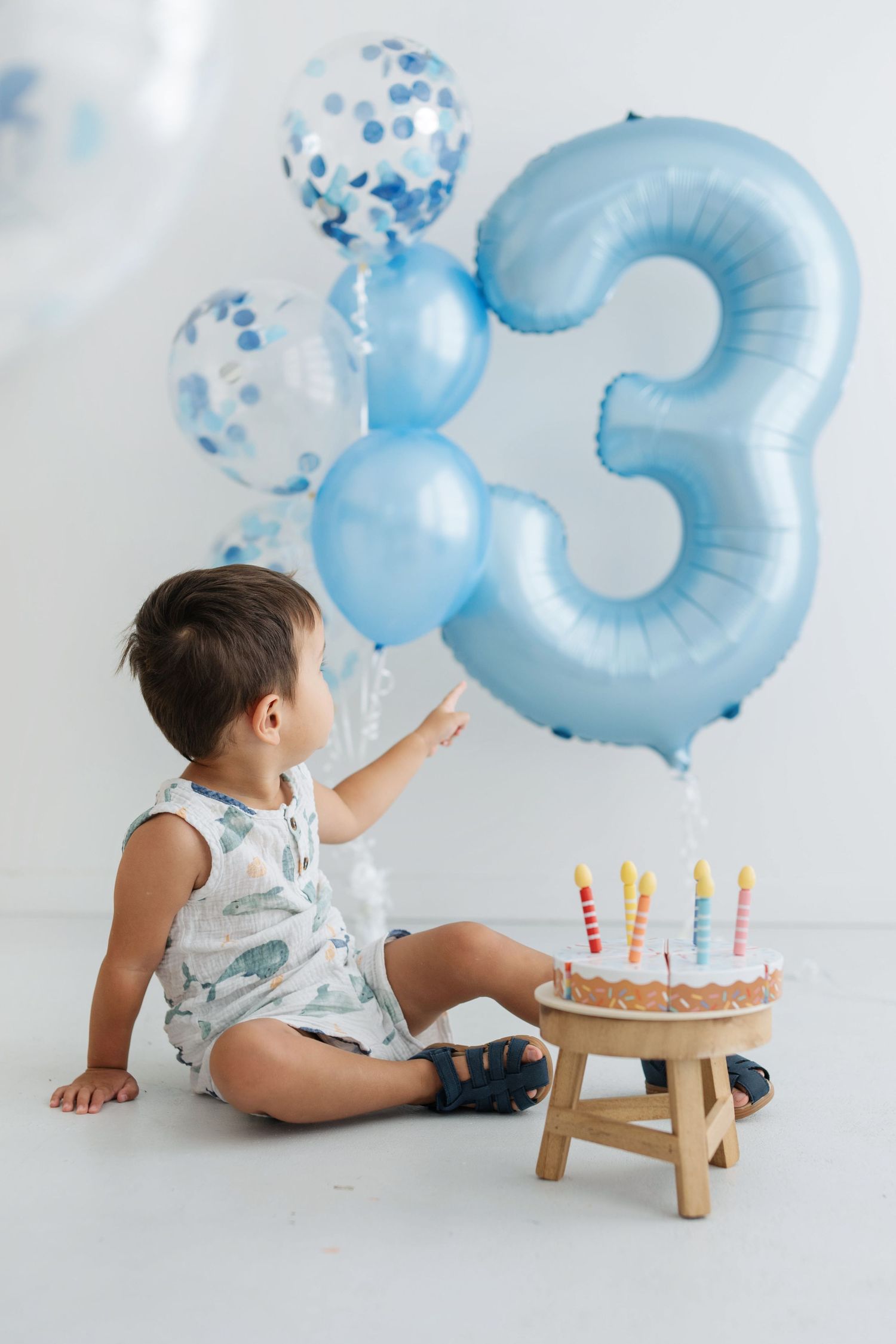 A toddler boy sits beside a birthday cake with candles and a large blue number 3 balloon on his 3rd birthday.