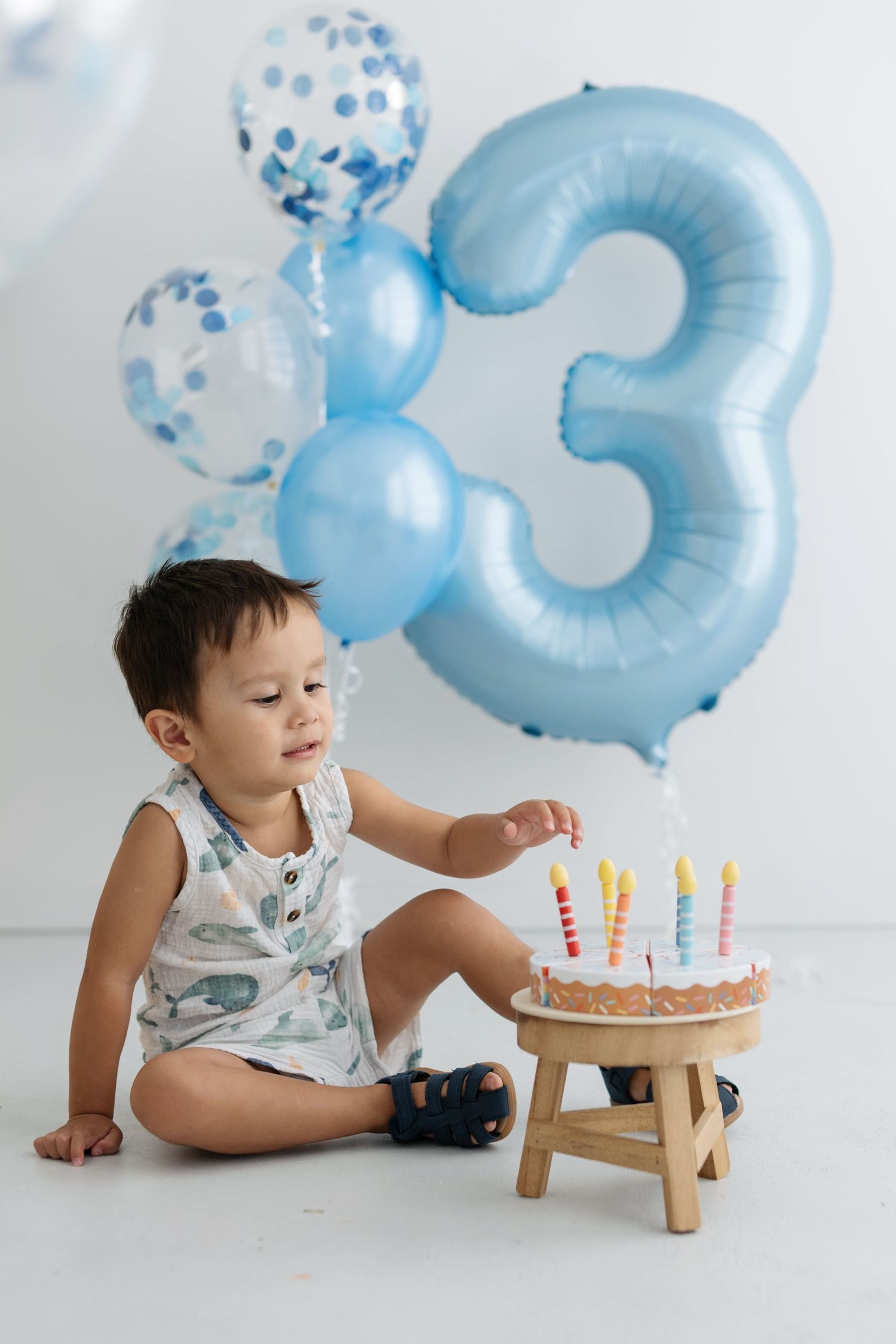 A toddler boy sits beside a birthday cake with candles, surrounded by blue balloons and a large number 3 balloon.