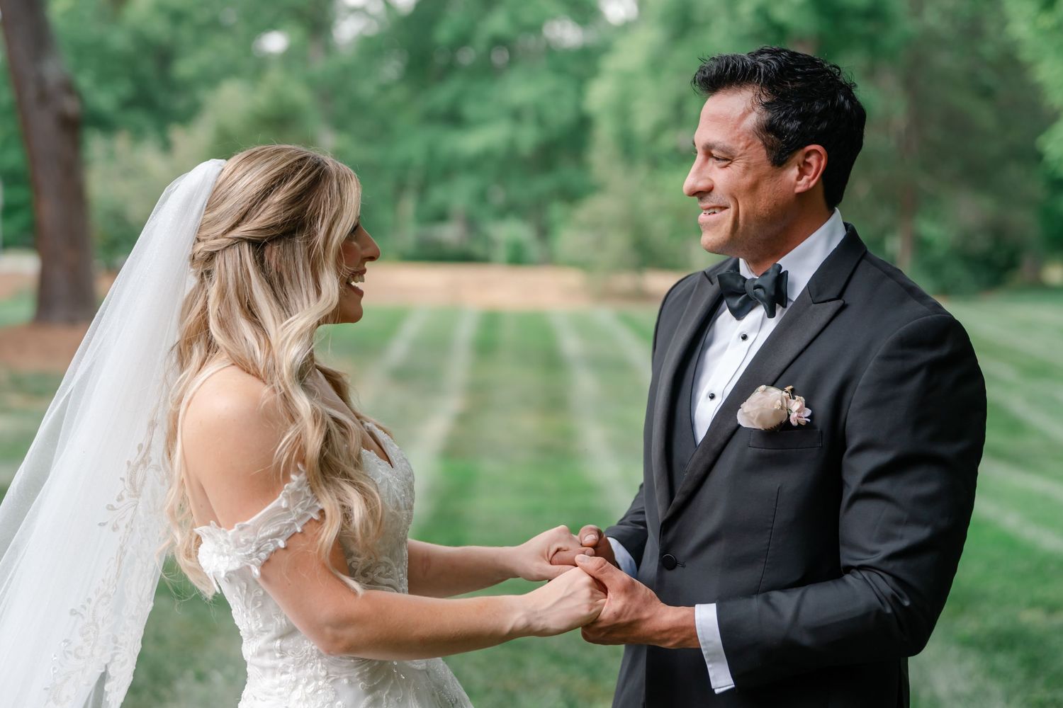 Bride and groom holding hands outdoors, smiling at each other in wedding attire with lush green trees in background.