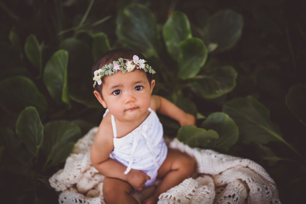 A cute baby girl wearing a floral crown sits in a white lace bowl surrounded by lush green leaves.