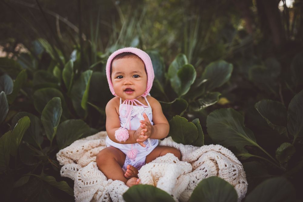 Adorable baby girl in pink bonnet and romper sitting on white blanket surrounded by lush green tropical plants.
