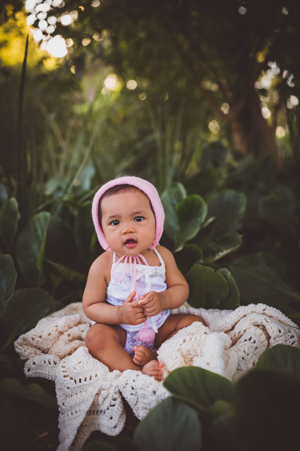 Adorable baby girl in pink bonnet and white outfit sitting on lace blanket surrounded by lush green garden foliage.