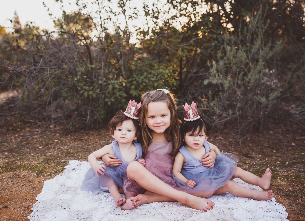 Three young girls wearing pink crowns and purple tutu dresses pose together on a white blanket outdoors.