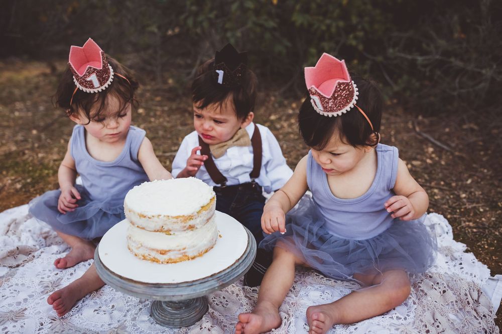 Three toddlers in gray tutu dresses and pink crowns sit outdoors around a white birthday cake on a silver stand.