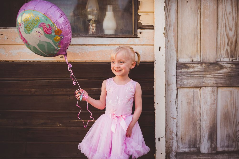 Smiling little girl in pink tutu dress holding a unicorn balloon, standing near rustic wooden doors outdoors.