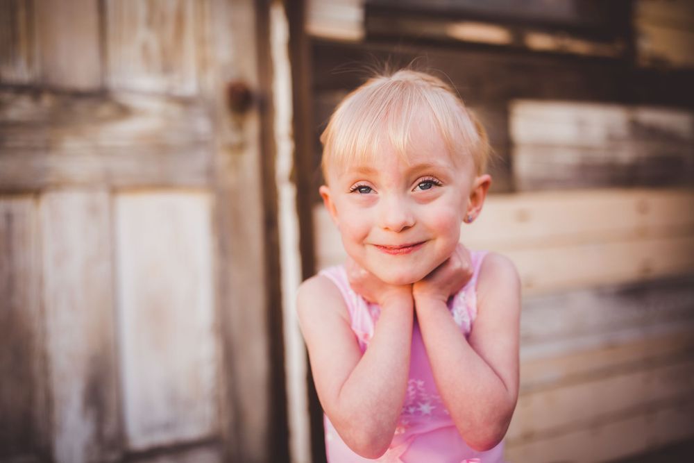 Smiling blonde toddler girl in pink dress posing with hands under chin near rustic wooden wall.