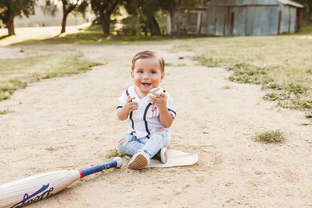 Smiling baby boy sitting on dirt path with cricket bat, wearing white outfit, enjoying outdoor photoshoot for his first birthday