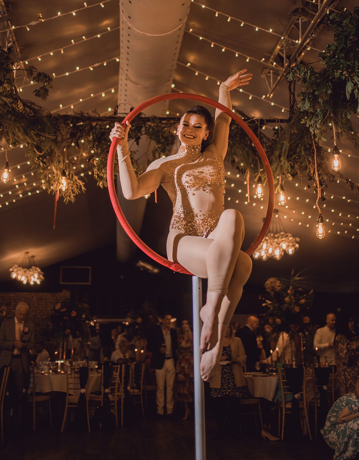 Aerial hoop performer in gold sequin leotard poses on lyra at elegant event venue with string lights.