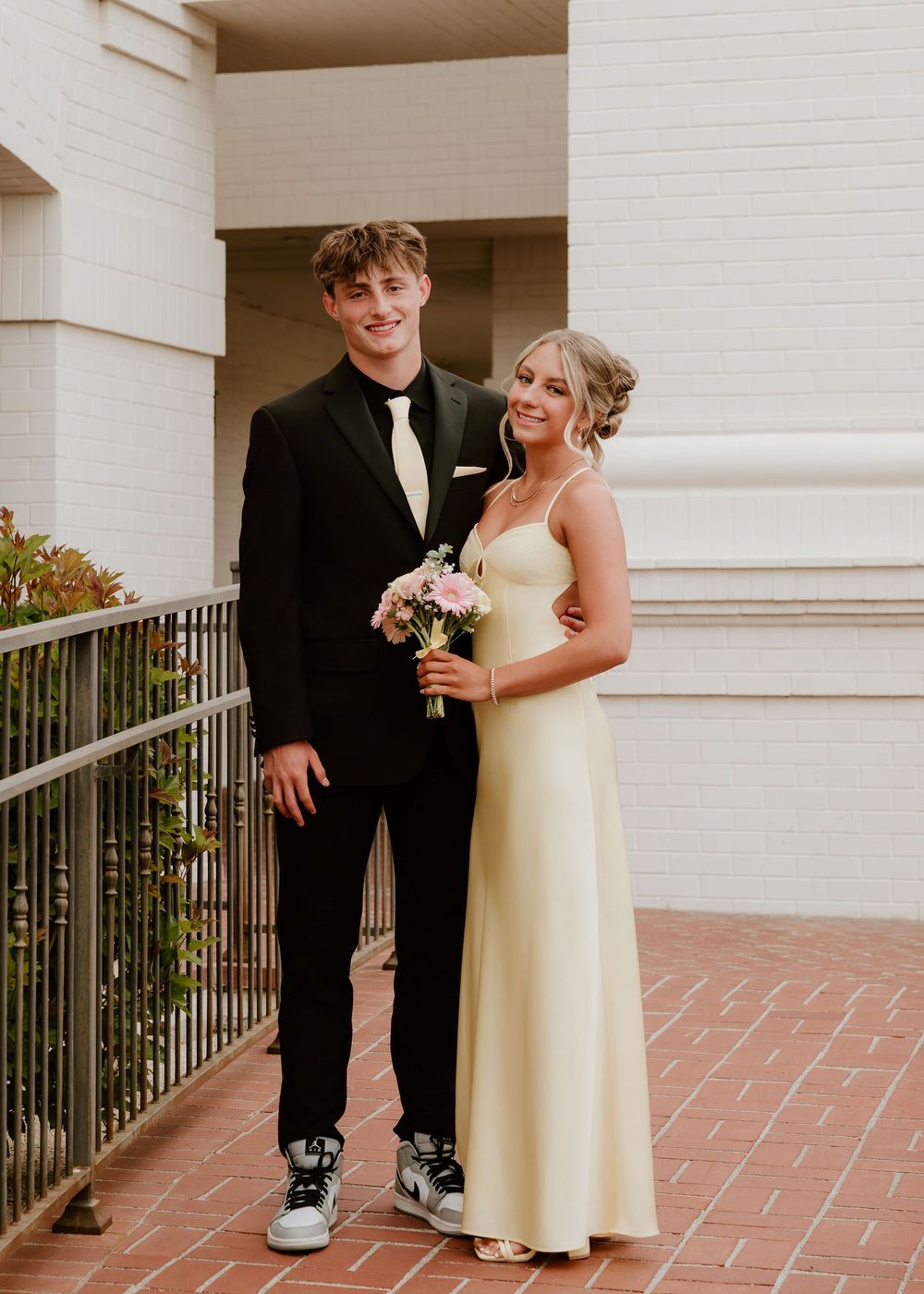 Young couple dressed for prom, man in black tuxedo and woman in yellow gown holding pink bouquet, posing outdoors.
