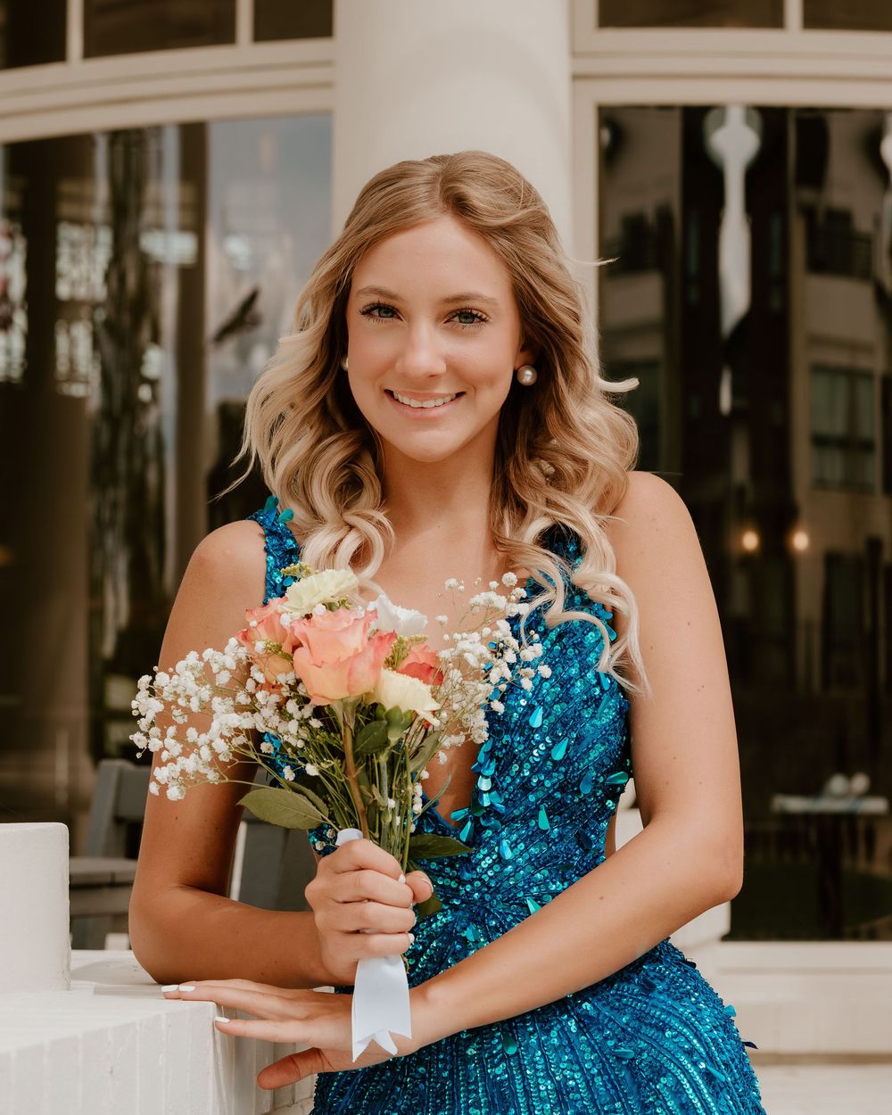 Young woman in blue sequin dress holding pink roses and baby's breath bouquet, smiling outdoors.