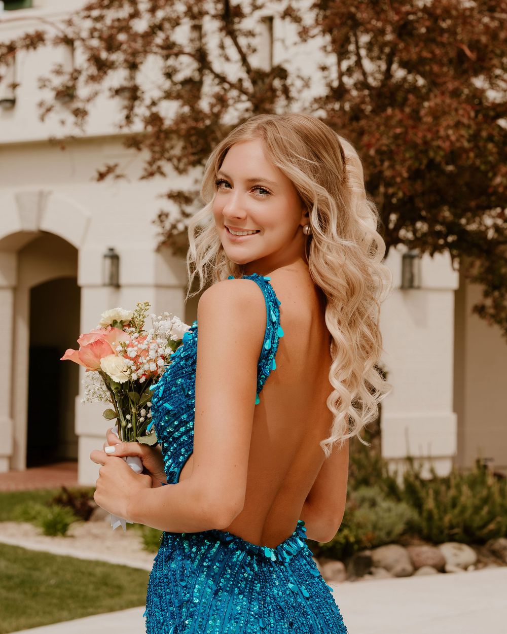 Young woman in backless teal sequin gown holding a bouquet, smiling over her shoulder outdoors.