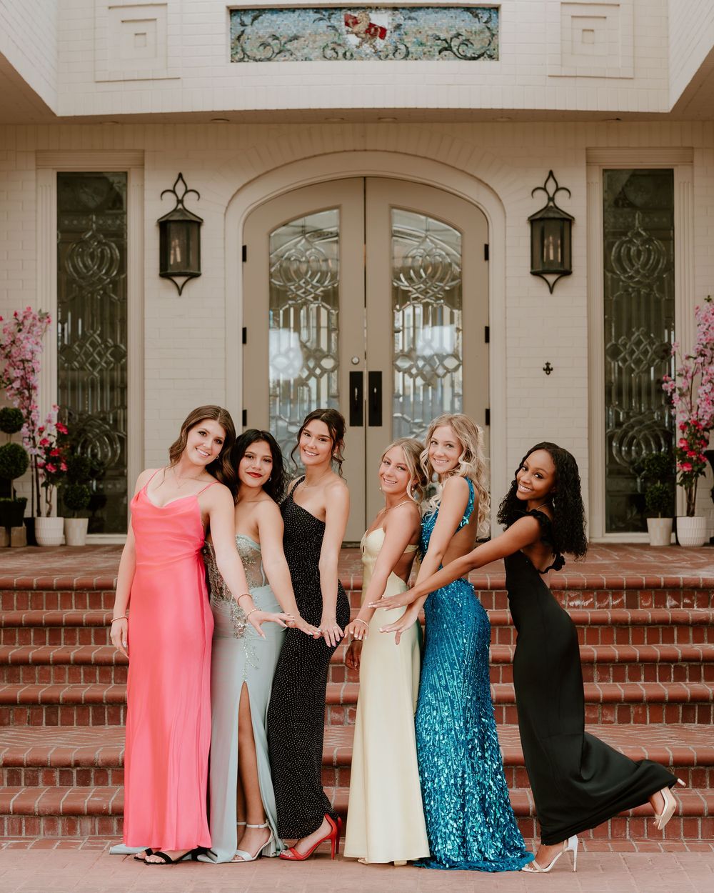 Six young women in elegant formal gowns pose together on brick steps outside a grand estate entrance.