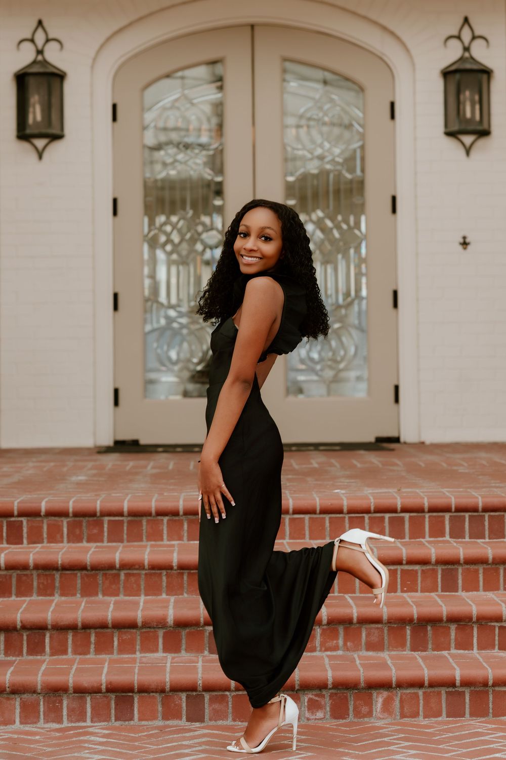 Woman in elegant black maxi dress and white heels posing on brick steps before ornate white double doors.