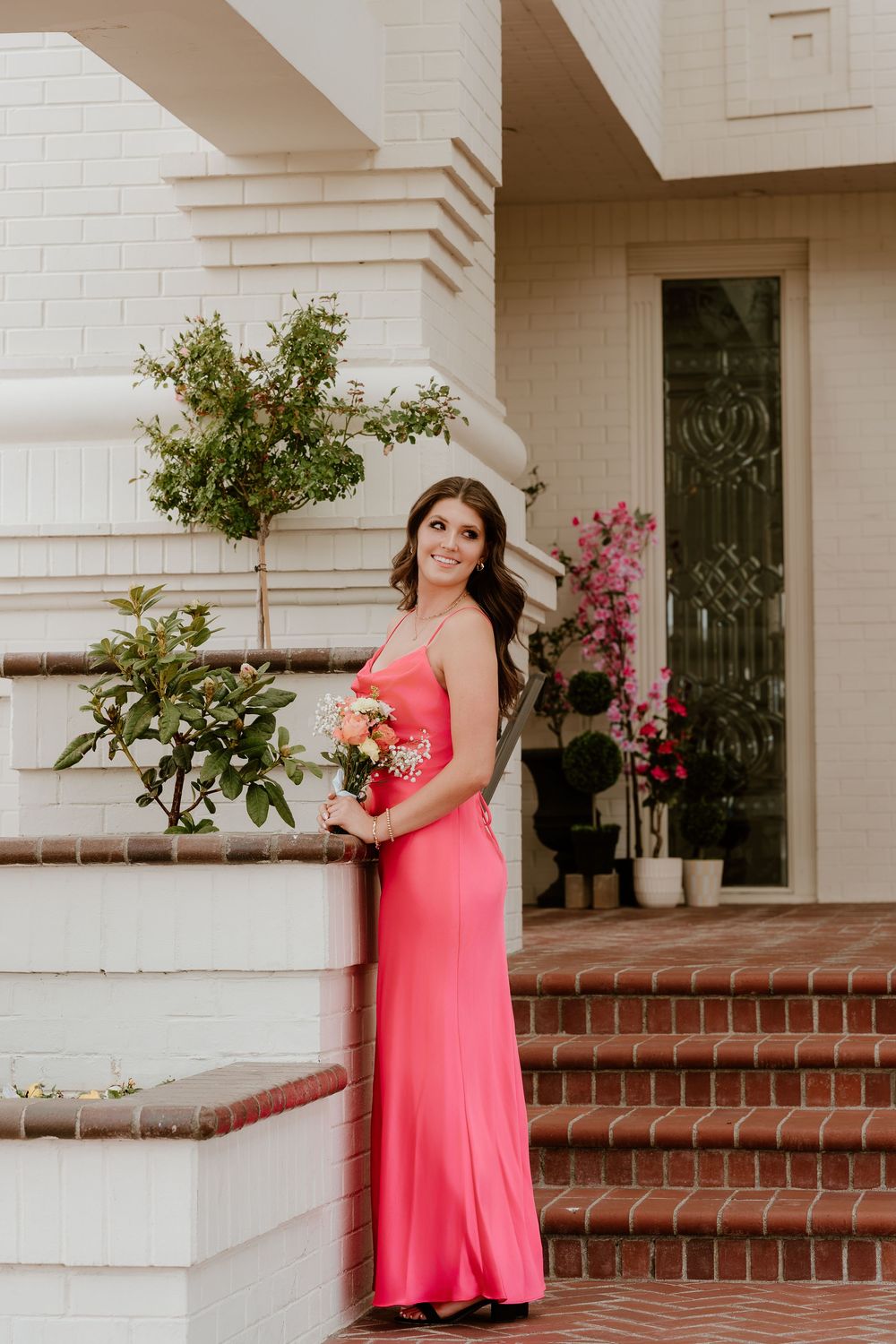 Woman in coral pink gown holding bouquet poses on brick steps outside white building with greenery.