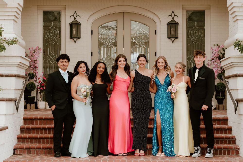 Group of teens in formal prom attire posing together on brick steps outside an elegant building.