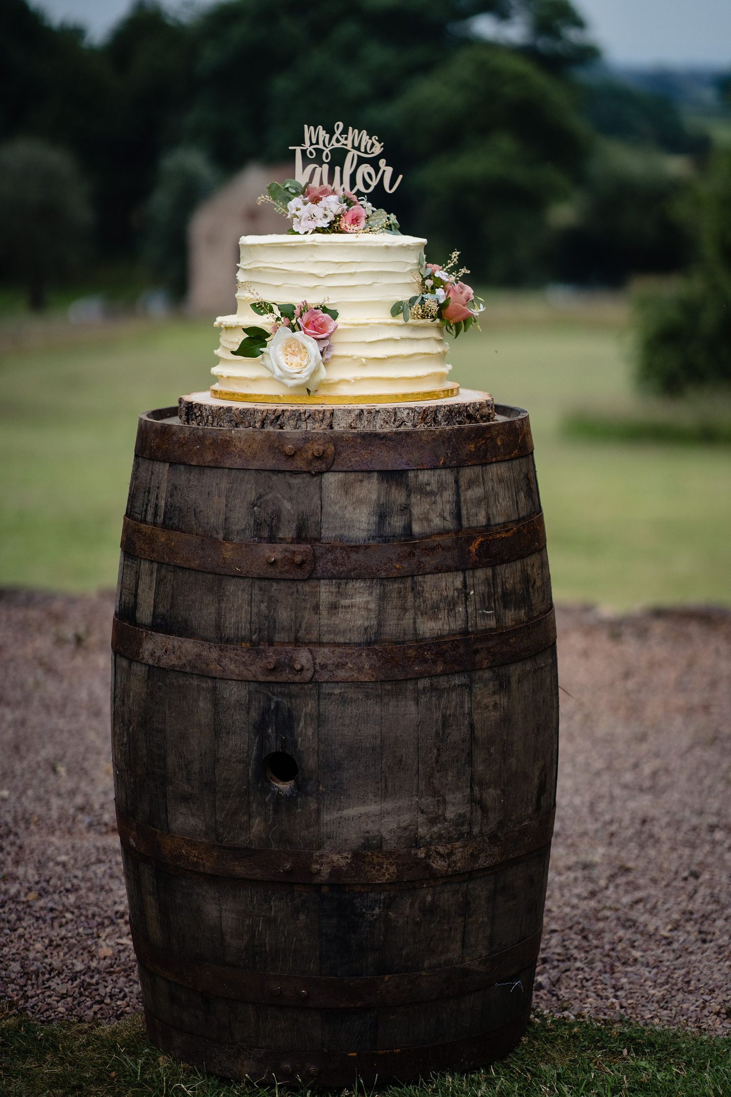 White wedding cake with floral decor and Mr & Mrs Taylor topper displayed on a rustic wooden barrel outdoors.