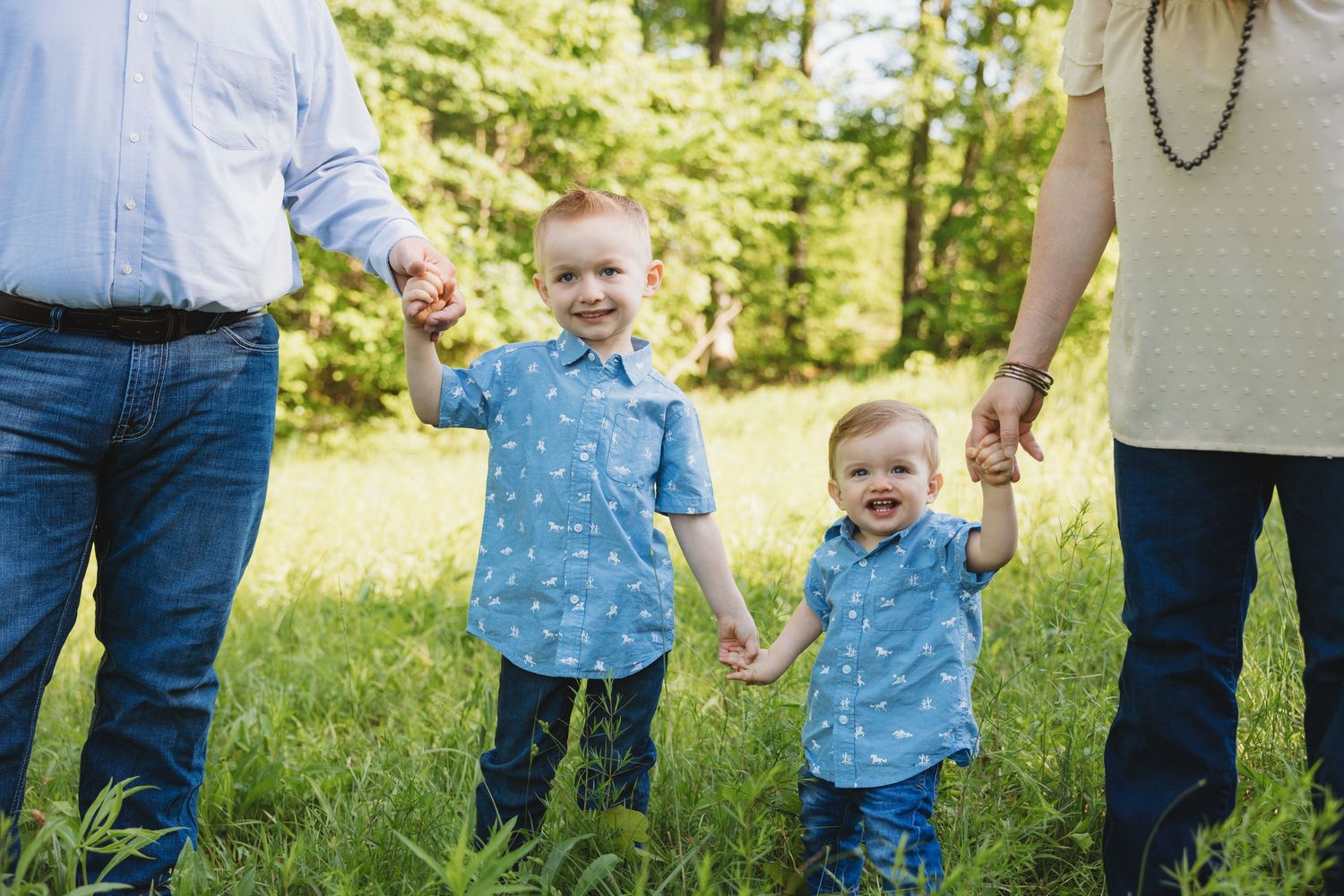 Two young boys in matching blue shirts hold hands with their parents in a sunny green field.