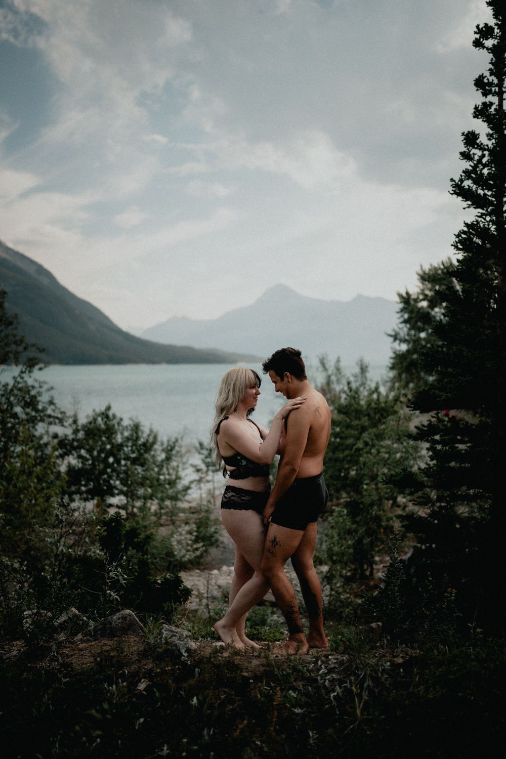 Couple in swimwear standing close together near mountain lake surrounded by trees.