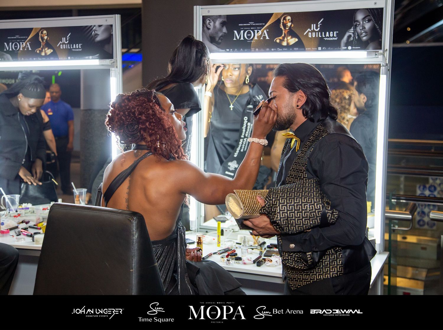 Makeup artist applying cosmetics backstage at MOPA event under bright vanity lights.