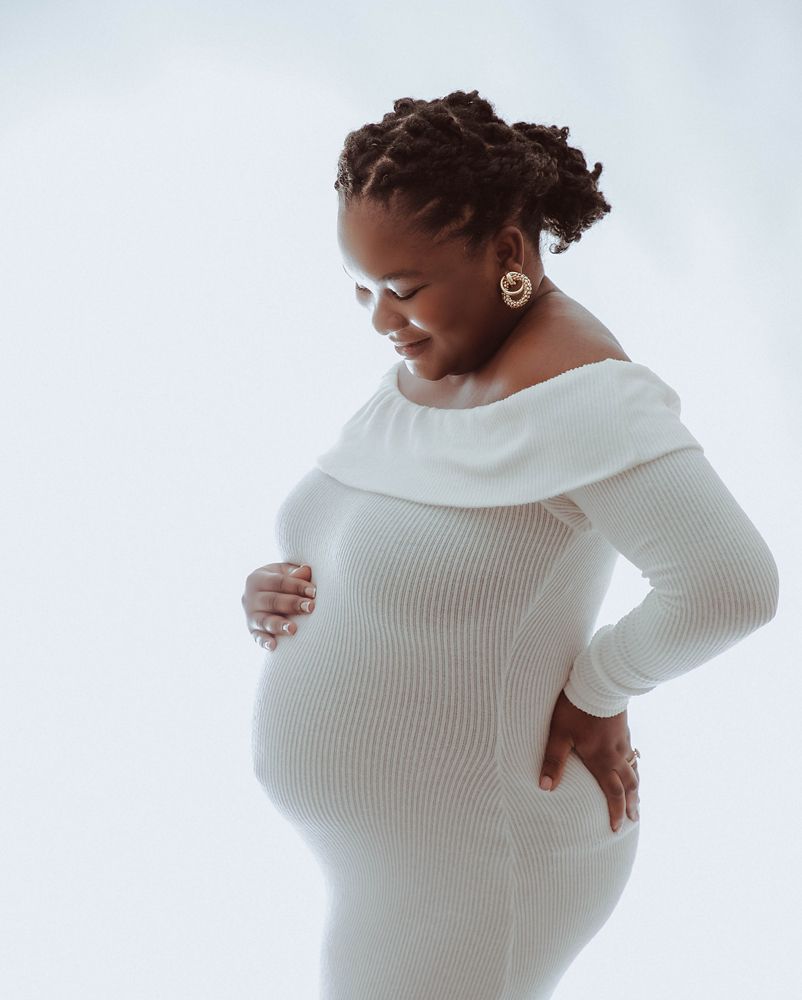 Pregnant woman in white off-shoulder ribbed dress cradling baby bump, smiling downward, studio maternity photo.