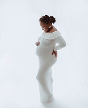 Pregnant woman in white off-shoulder maternity gown cradling baby bump during elegant studio maternity photoshoot.