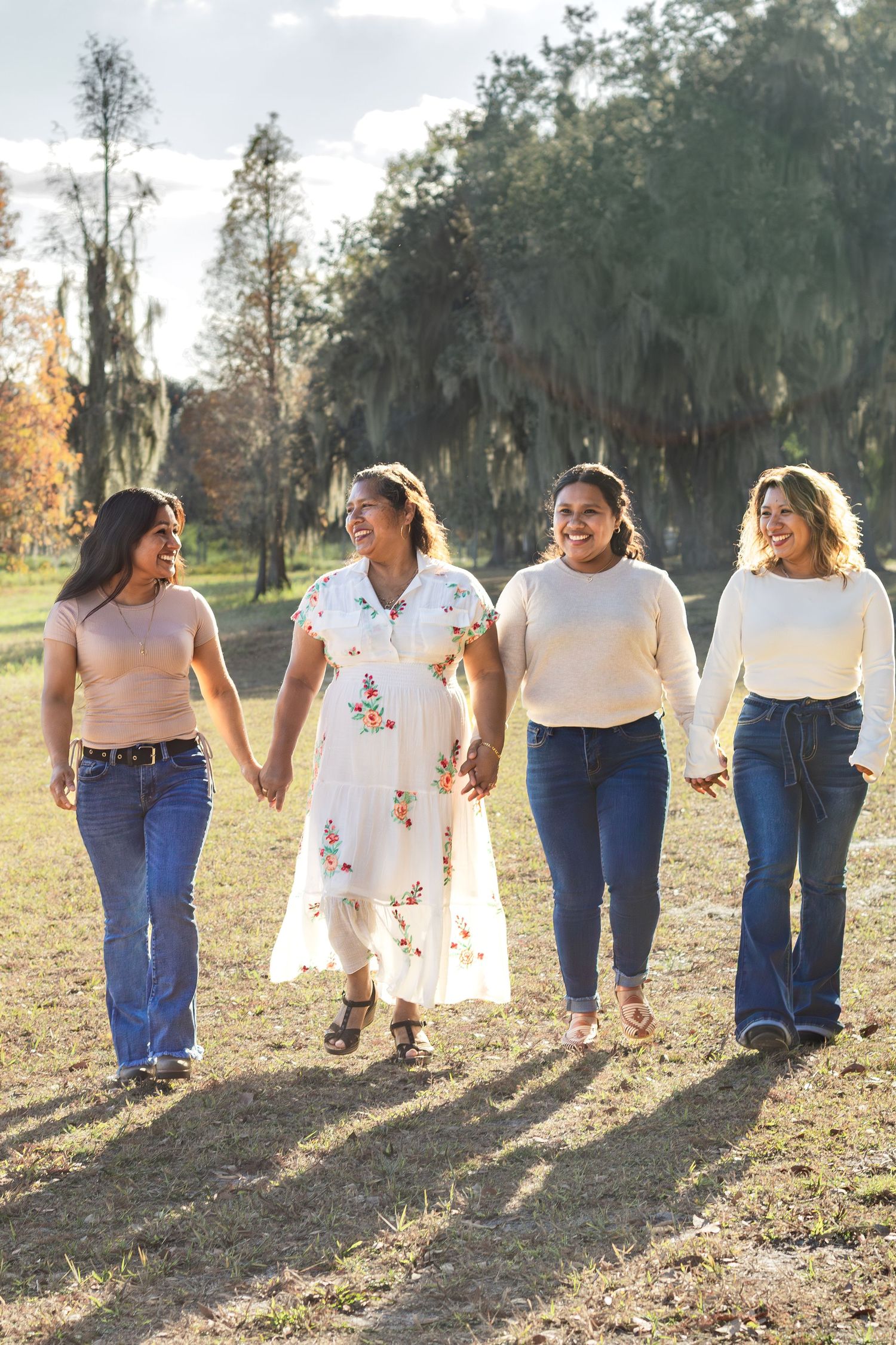 Four joyful women holding hands and walking together through a sunny autumn park setting.