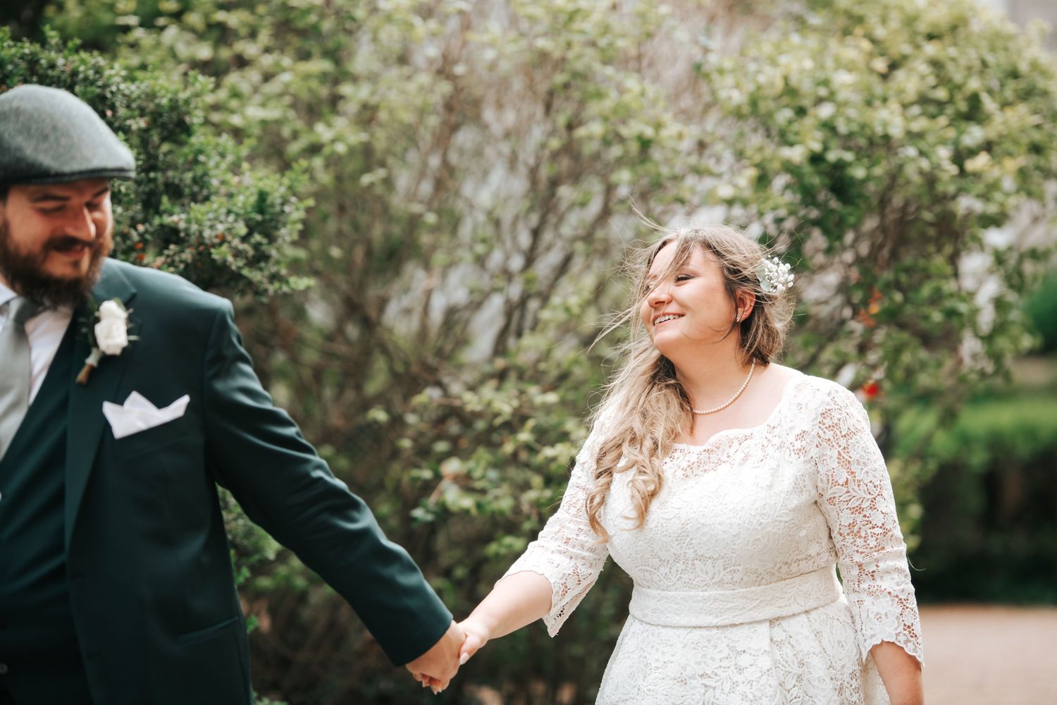 Happy bride in lace dress holding groom's hand, laughing outdoors among green trees.