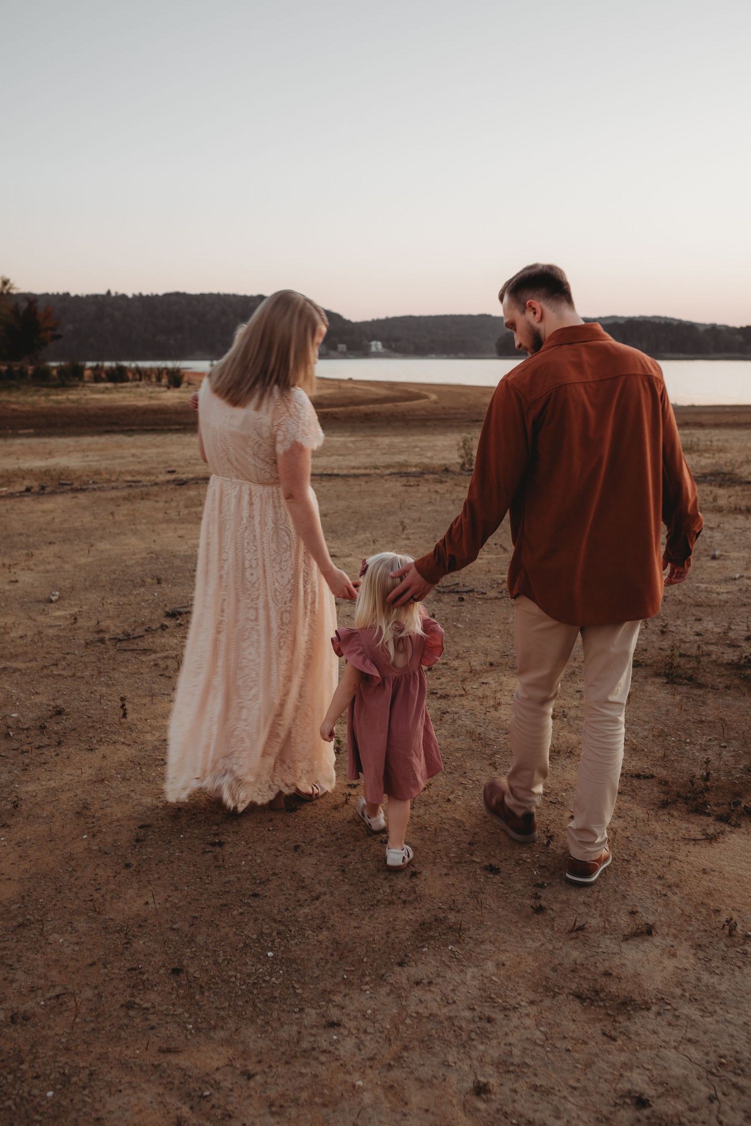 Family of three holding hands walking toward a serene lake at sunset, mother in white dress, father in rust shirt.