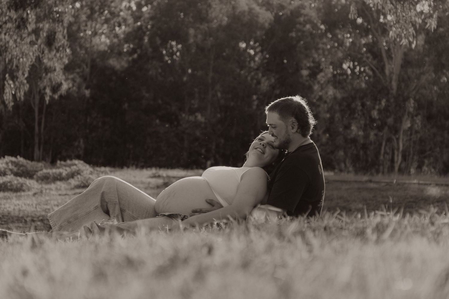 Black and white photo of a pregnant couple relaxing together on grass in a park.