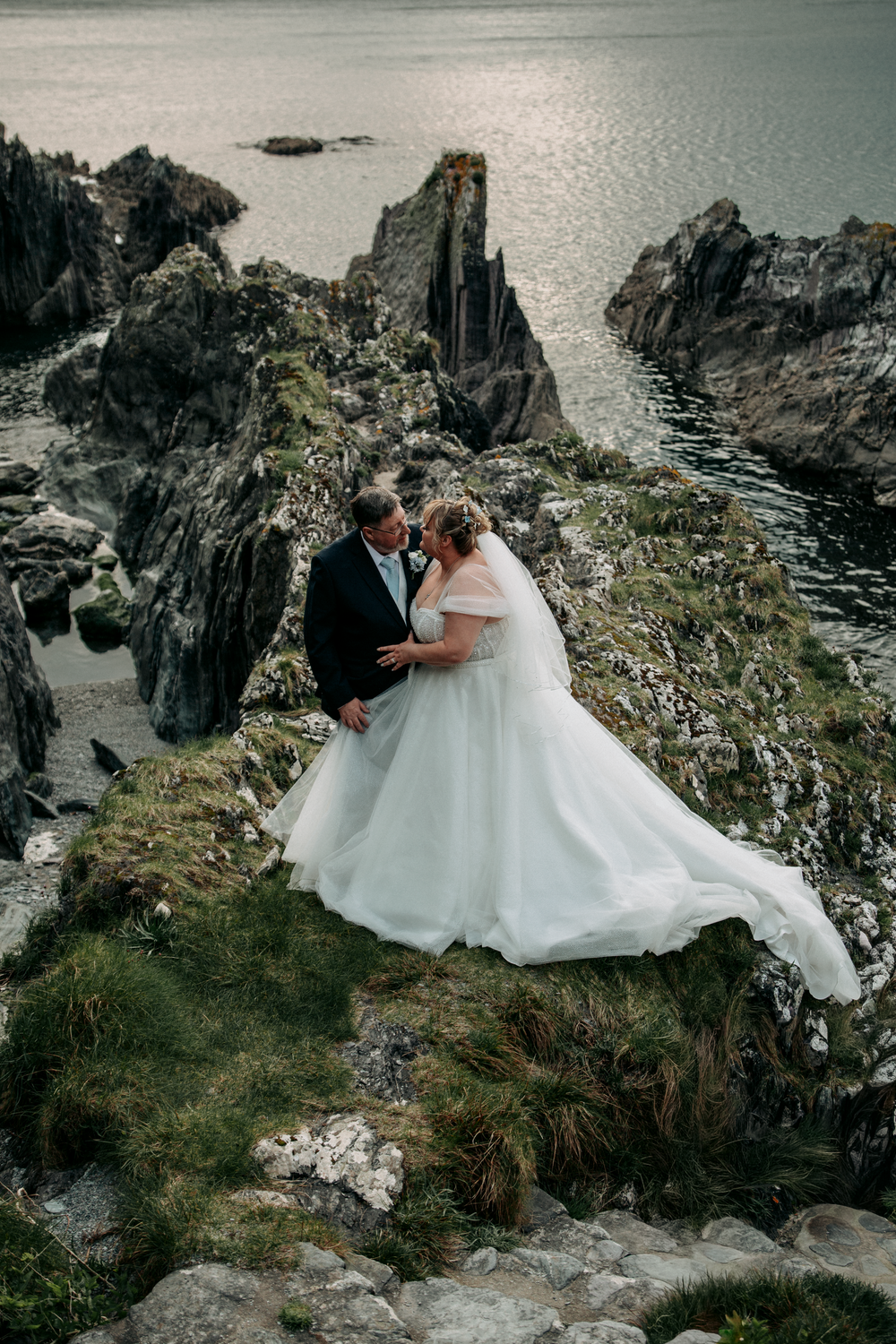 Bride and groom embrace on dramatic rocky coastal cliffs, white gown flowing in the wind, ocean backdrop.
