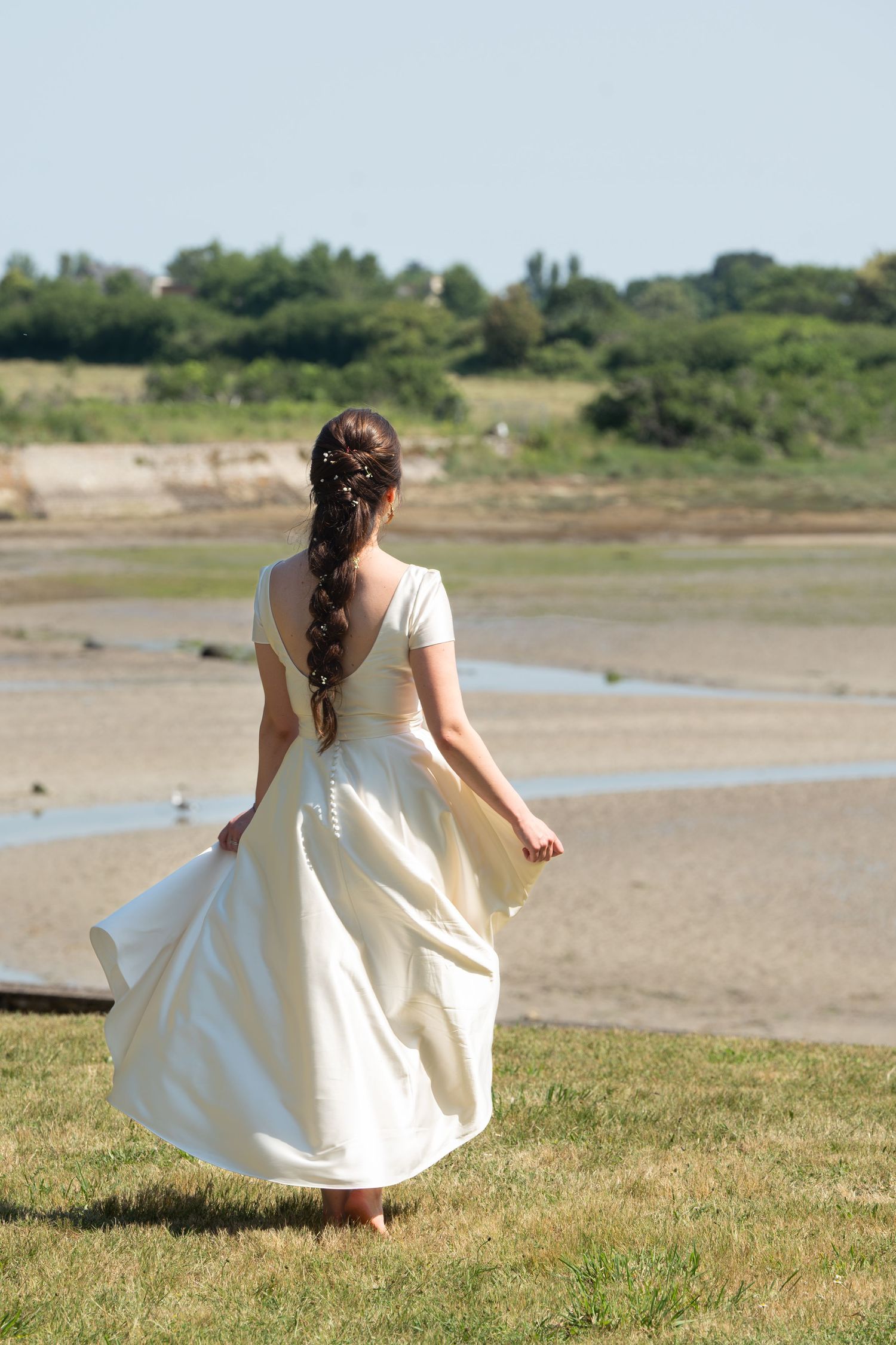 Une mariée vêtue d'une robe de mariée blanche mi-longue, marchant pieds nus dans une prairie côtière, vue de dos. photo lecaude stephanie