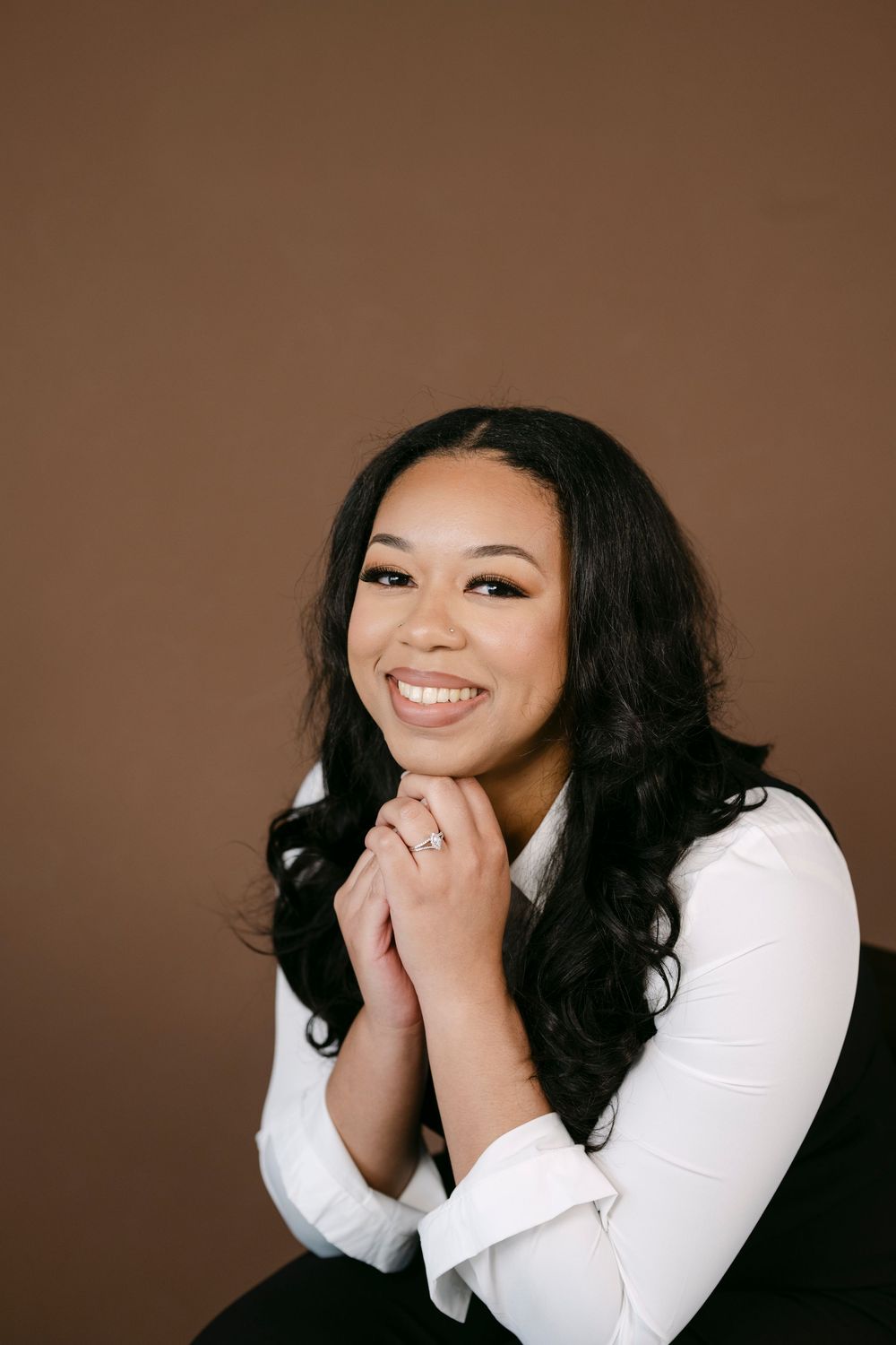 Smiling young woman in white blouse posing with hands clasped under chin against brown studio background.