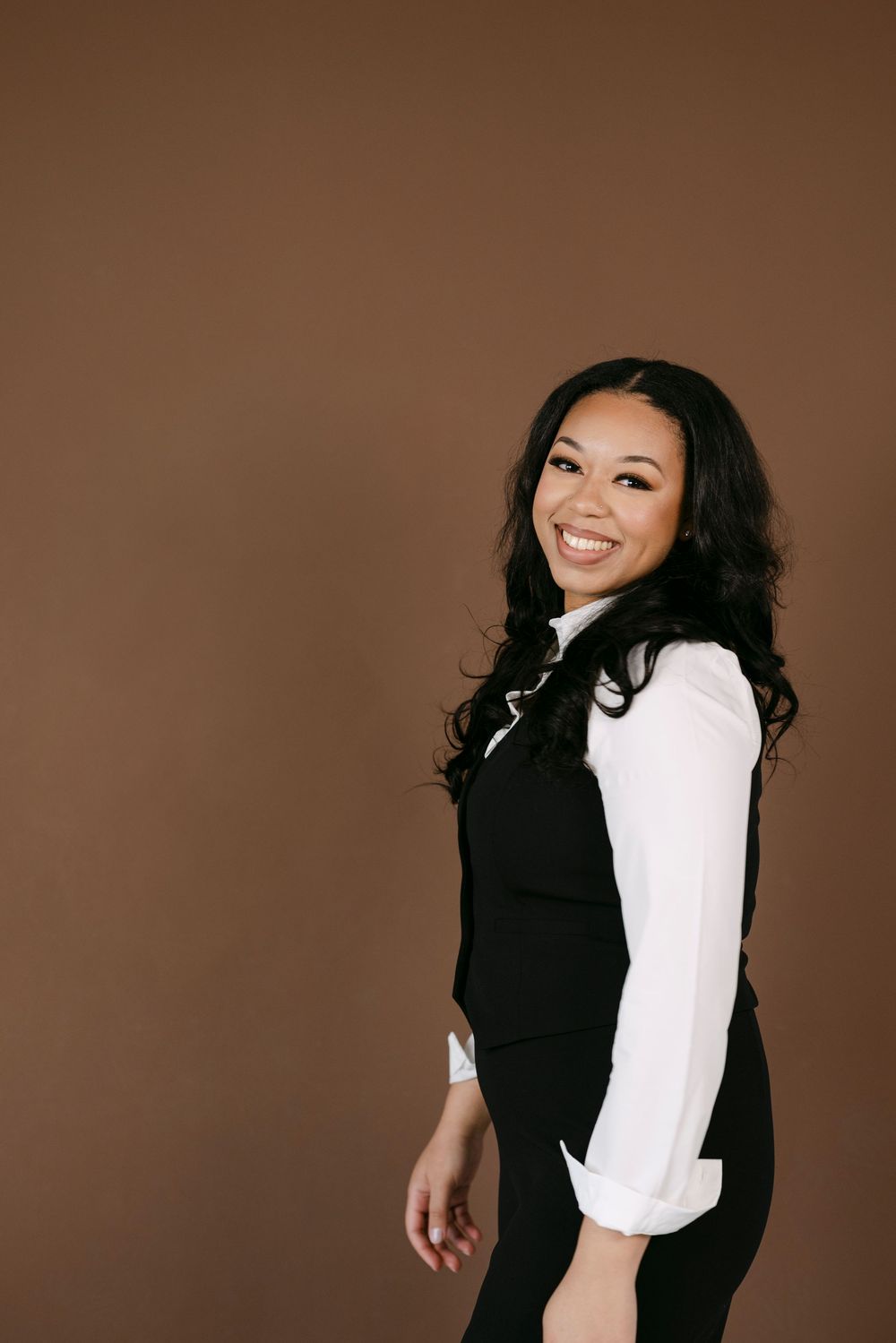 Smiling young woman in black vest and white shirt posing against a brown background.