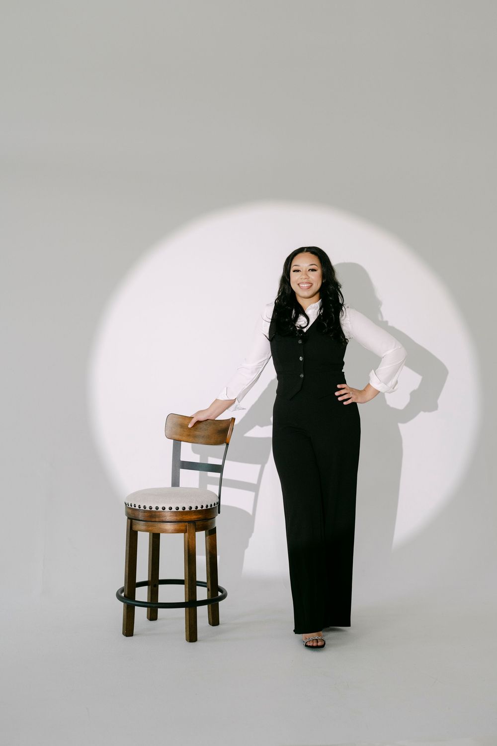 Woman in black vest and wide-leg pants poses beside wooden stool under spotlight in professional studio photo.
