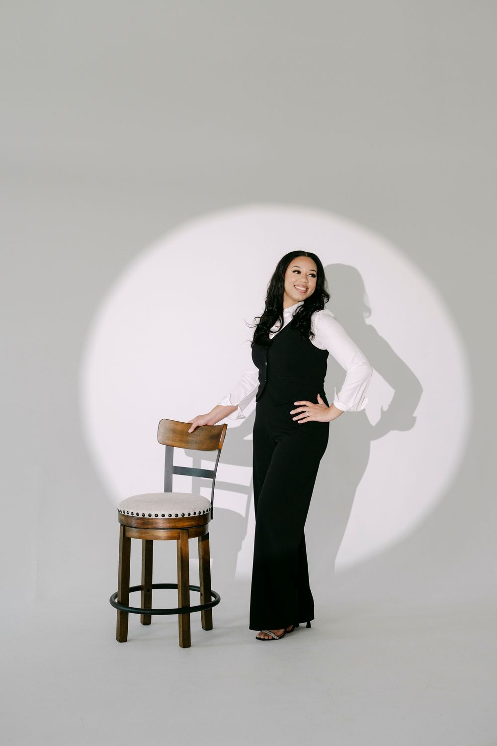 Woman in black vest and pants poses confidently beside a wooden stool under a spotlight on white background.