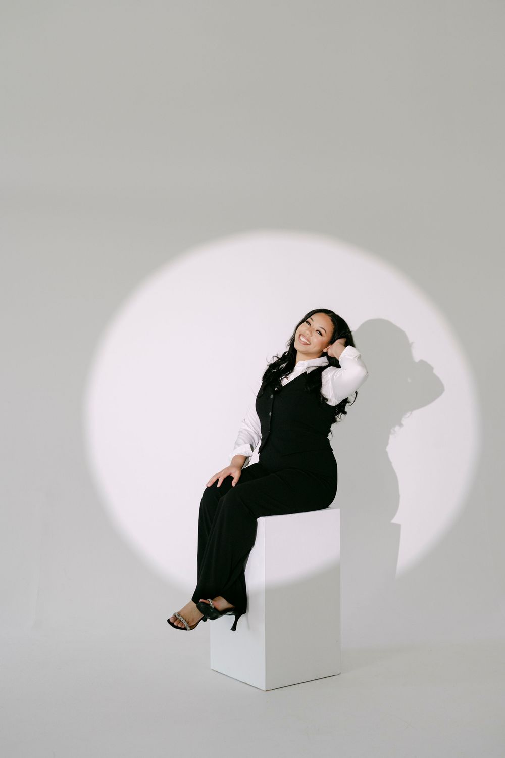 Woman in black vest and trousers sits on white cube under spotlight with dramatic shadow on gray background.