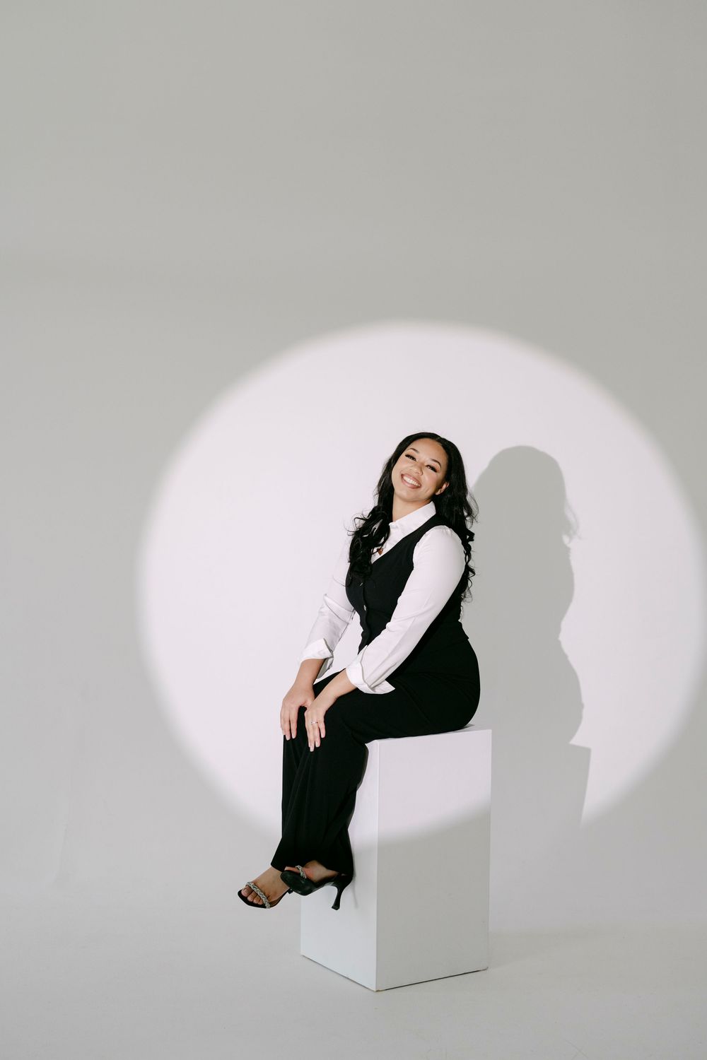 Woman in black vest and white blouse sits on white cube under spotlight against gray background, smiling confidently.