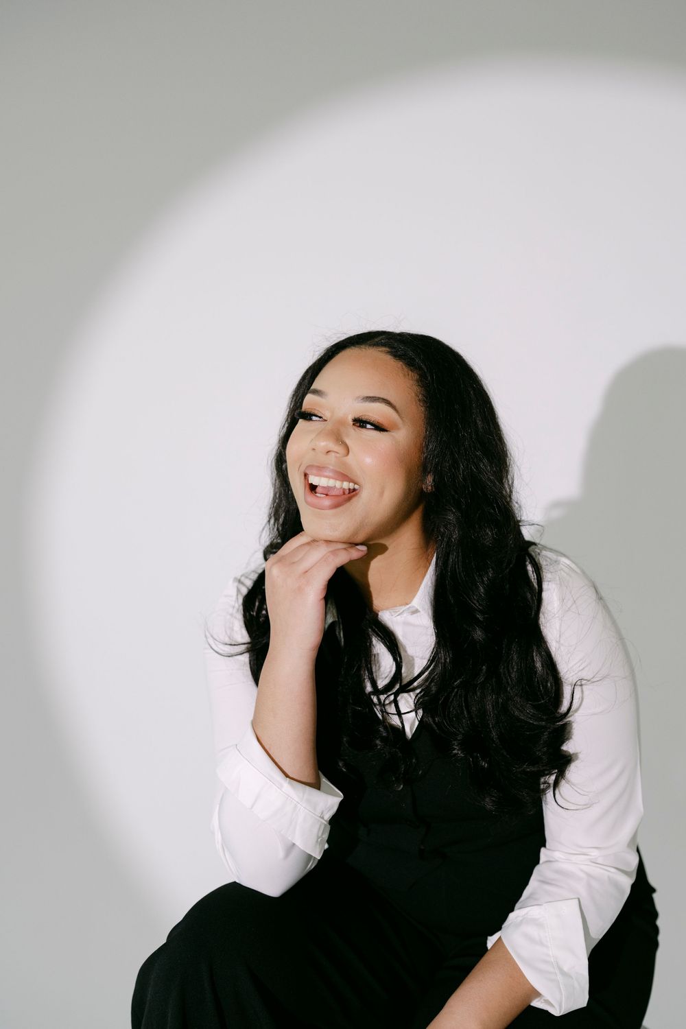 Young woman with long black hair smiling, wearing white shirt and black overalls, posing against gray background.