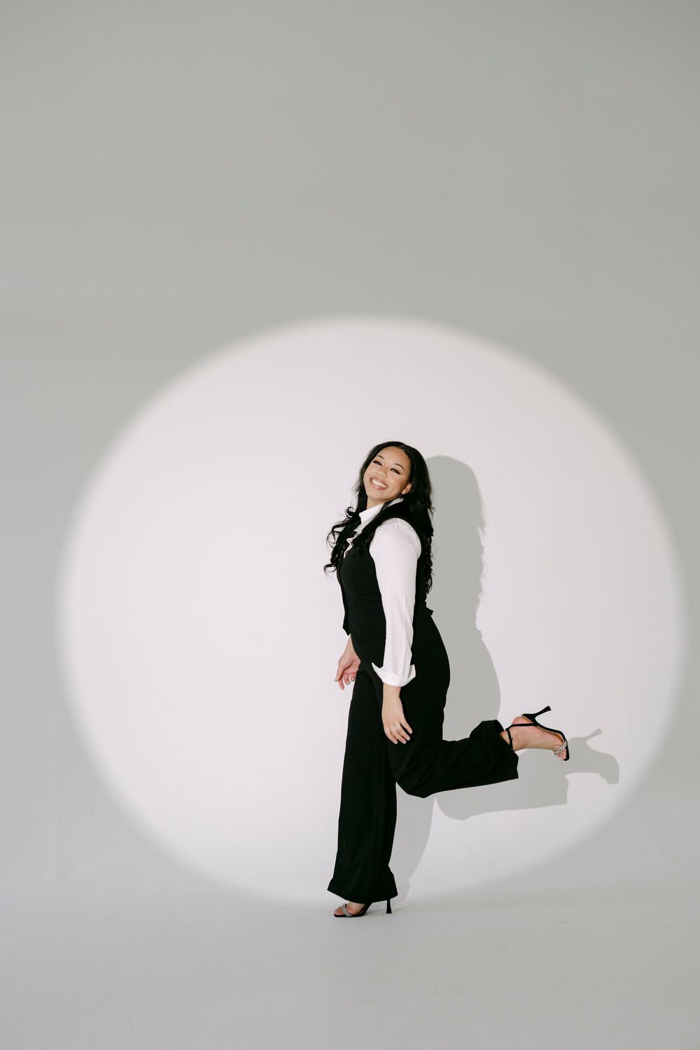 A stylish woman in black vest and trousers with heels poses confidently under a spotlight against a gray background.