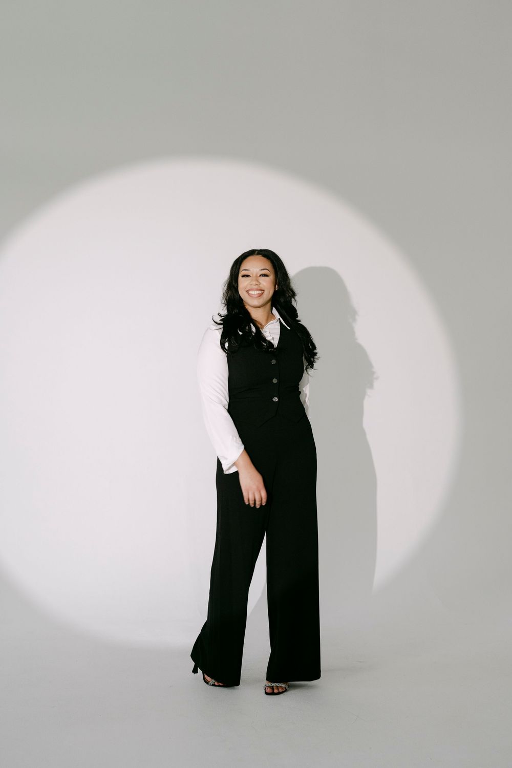 A young woman in a black vest and wide-leg pants poses confidently under a spotlight against a gray background.