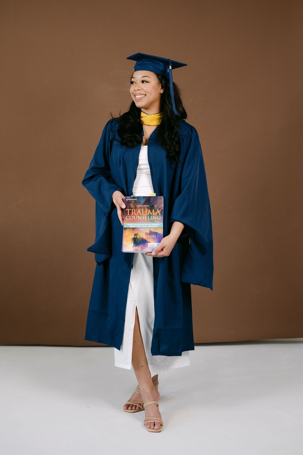 Smiling woman in navy blue graduation cap and gown holding a book, posing against a brown background.