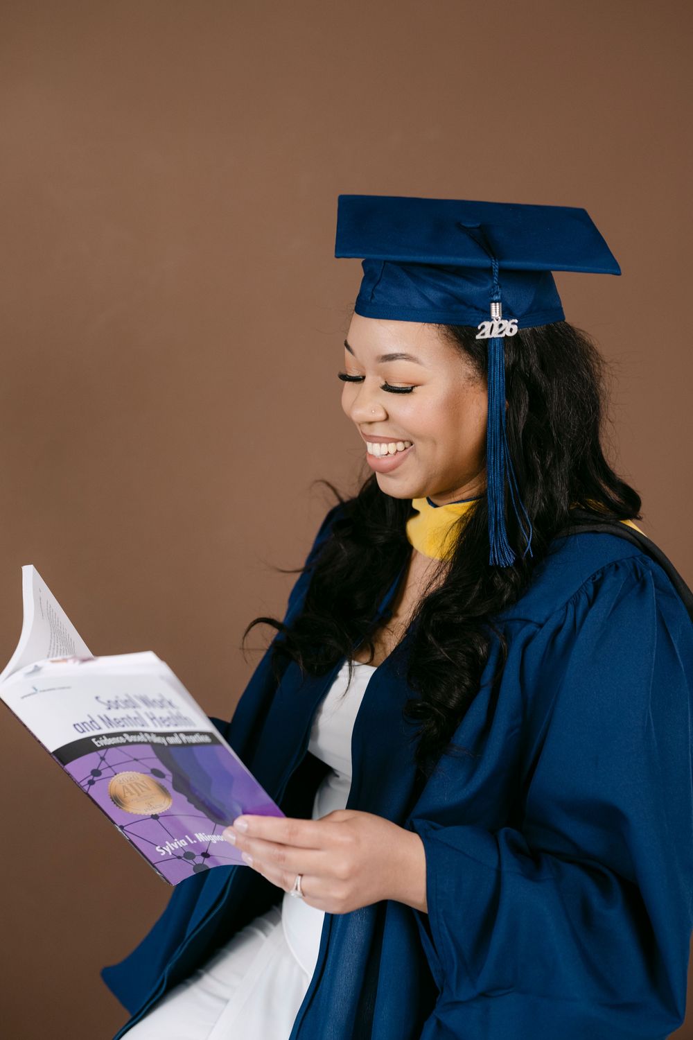 Smiling graduate in blue cap and gown reading a book, wearing a 2026 graduation cap charm.
