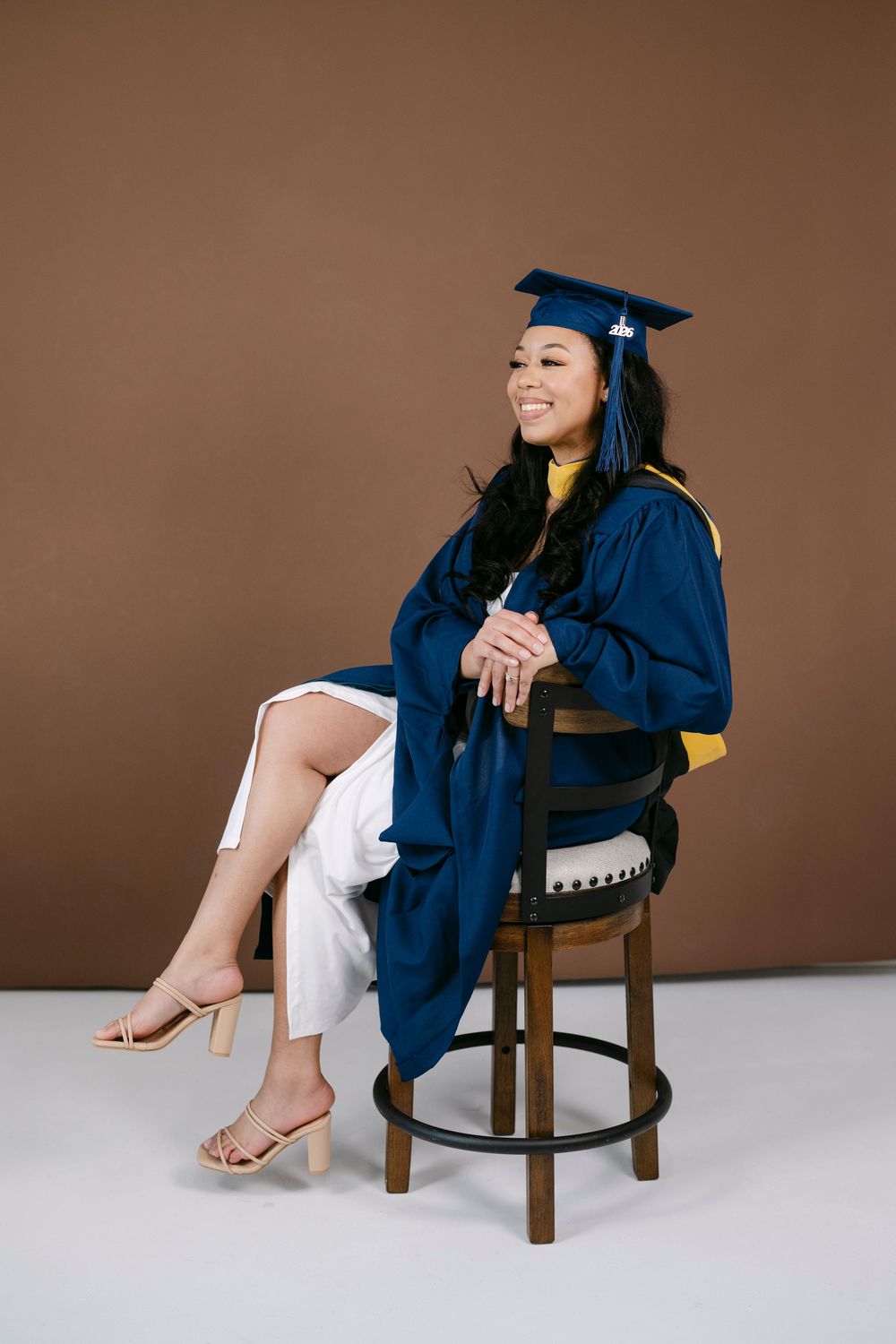 Happy graduate in blue cap and gown sitting on chair against brown studio background wearing white pants.