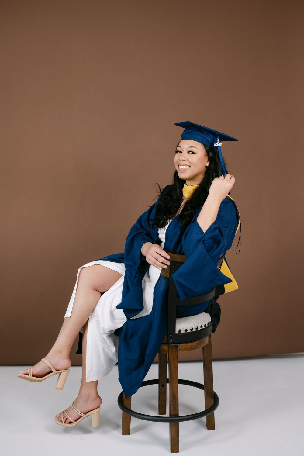 Smiling woman in blue graduation cap and gown seated on a stool against a brown background.