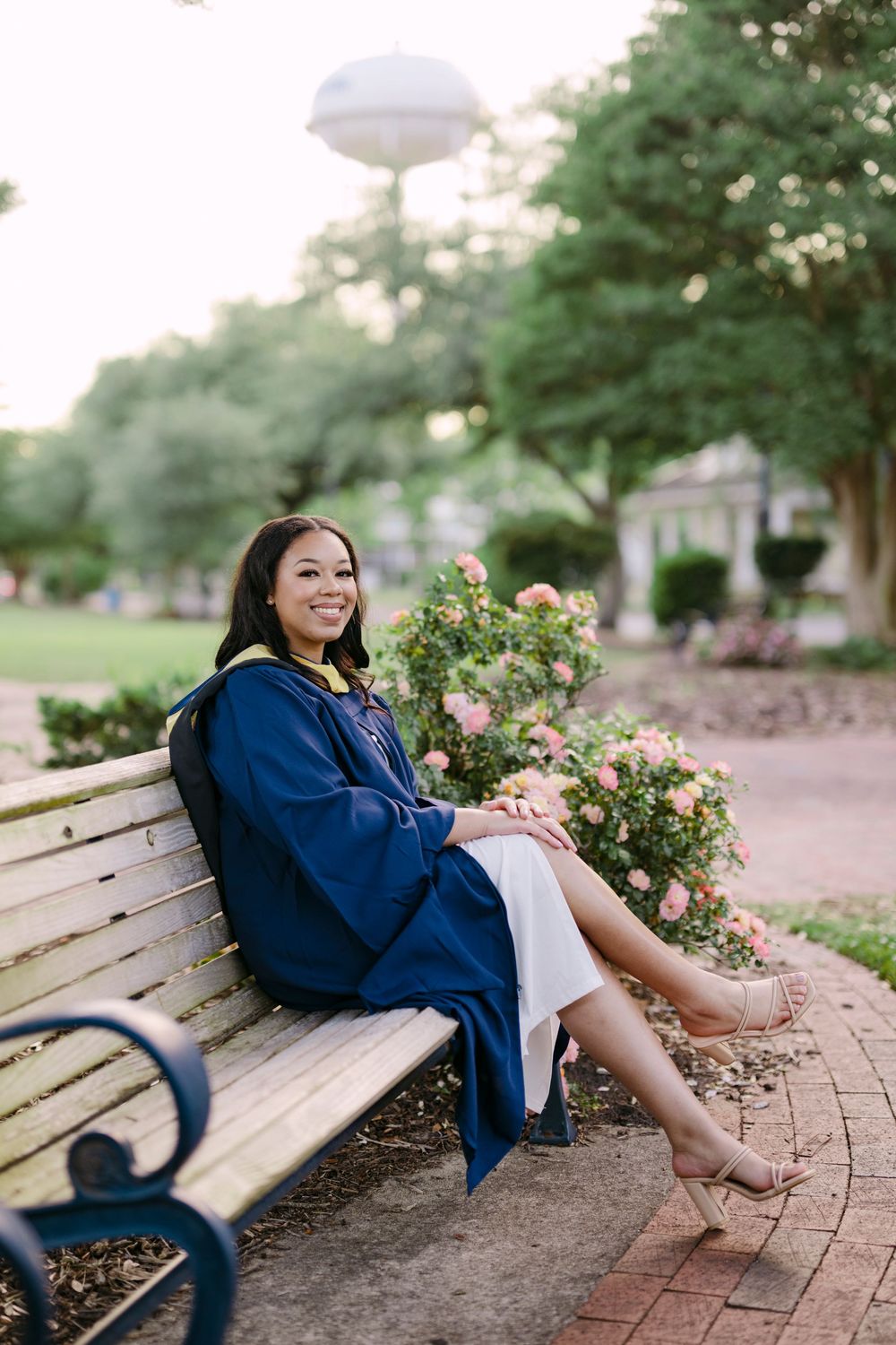 A smiling graduate in a navy blue cap and gown sits on a park bench surrounded by blooming pink roses.