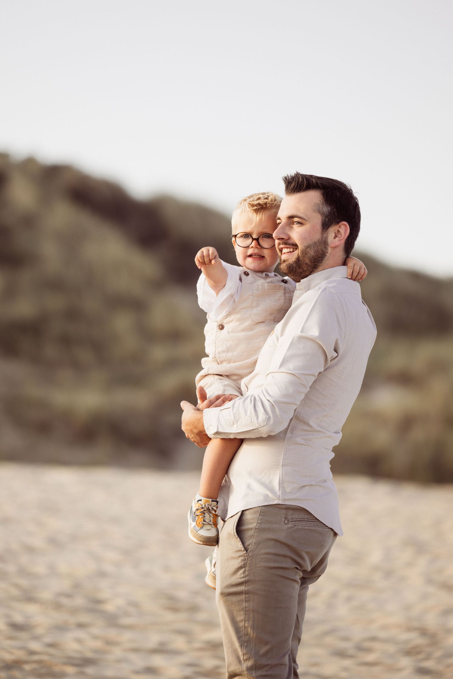 A parent and child share tender moments together on a sandy beach in casual white and beige attire.