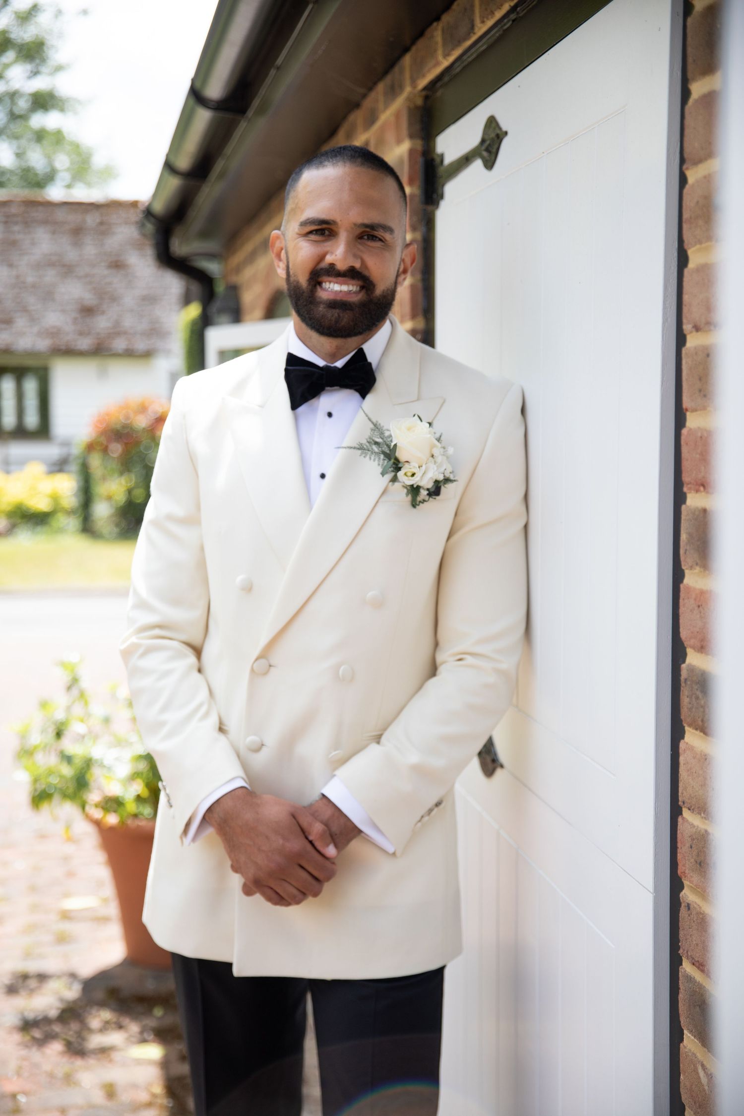 A groom in a cream double-breasted tuxedo jacket with black bow tie and white boutonniere poses outside a brick building.