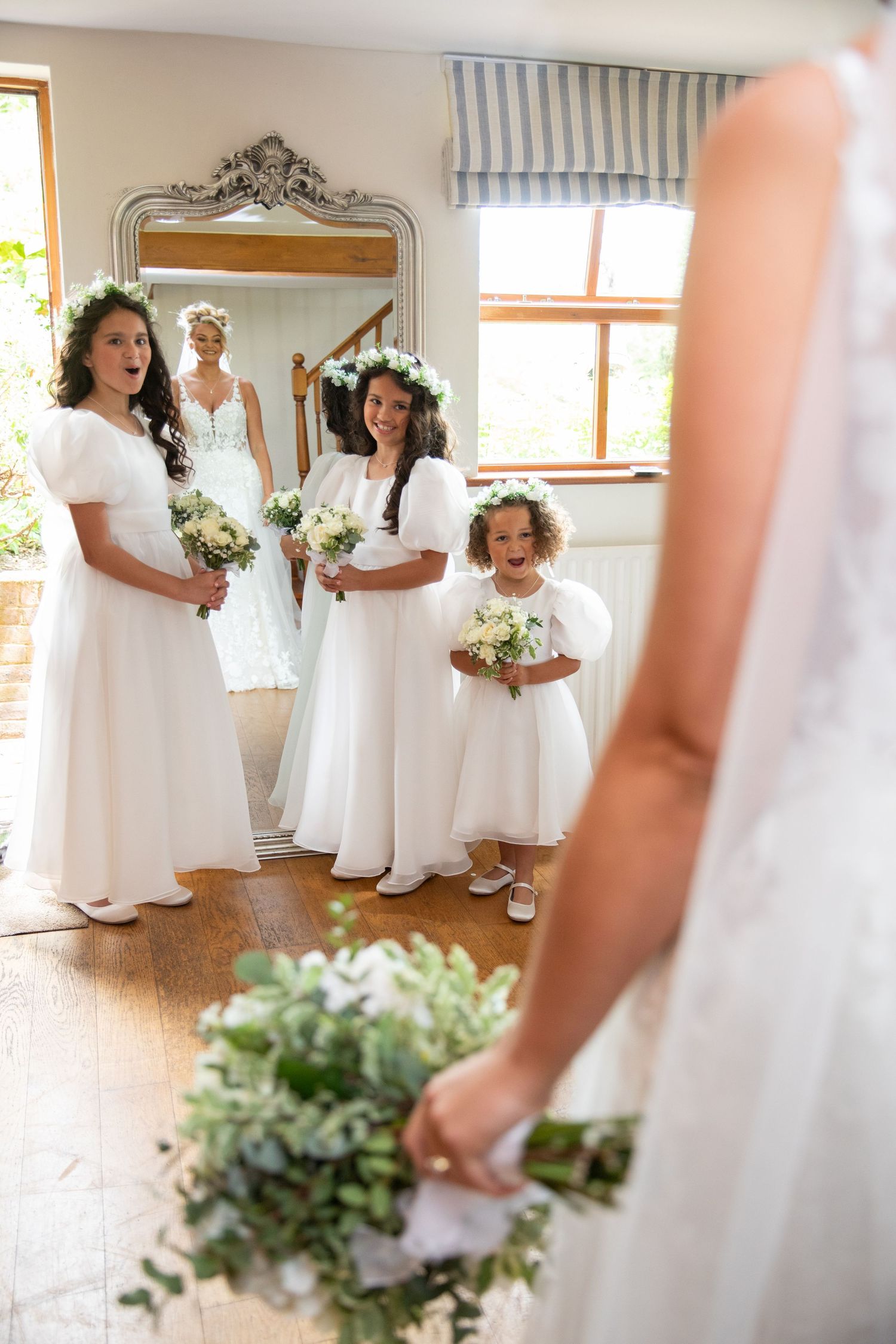 Bridesmaids in flowing white dresses hold elegant bouquets while posing in a sunlit room during wedding preparations.
